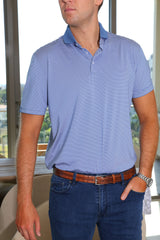 A man in a blue and white striped polo shirt stands with his arm relaxed at his side and a wooden table in the background.