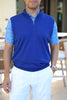 A man wearing a blue sleeveless pullover and light blue patterned shirt stands in a sunny outdoor setting.