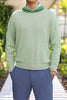 A man in a light green sweater with a hood and dark shorts stands with his hands in his pockets against a blurred green backdrop.
