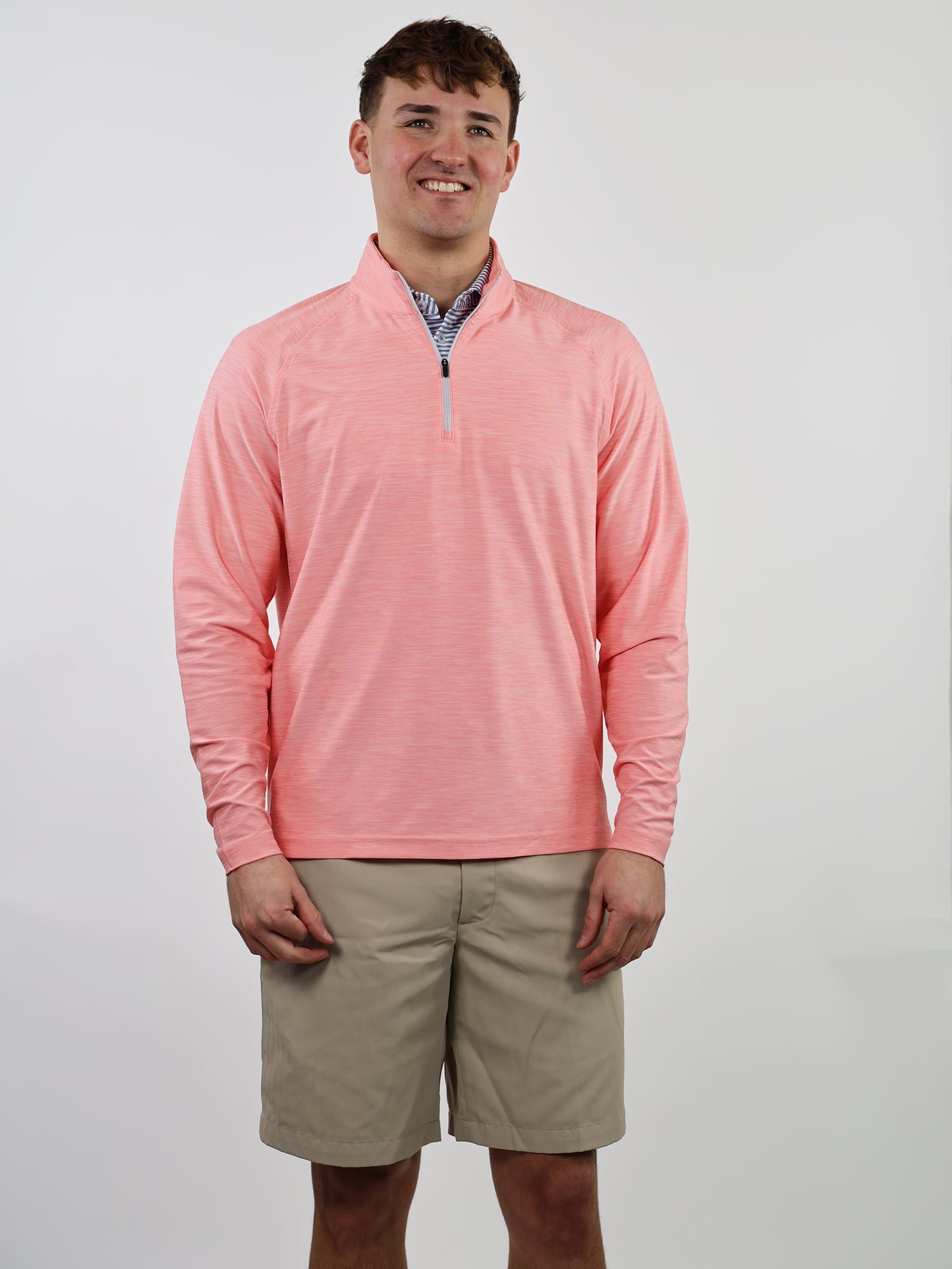 A man in a coral long-sleeve, quarter-zip top and beige shorts smiles against a plain white background