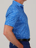 Man in a side profile wears a blue patterned short-sleeve polo shirt with floral designs, paired with dark pants and a black belt against a neutral background.