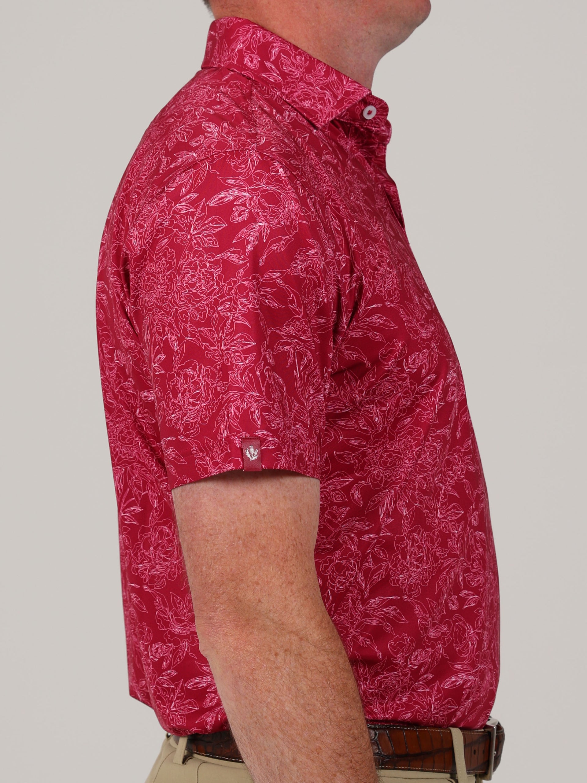 A man in a red short-sleeved shirt with a white floral pattern is shown from the side. He wears tan pants and a brown belt against a neutral background.