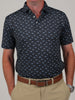 Man wearing a navy polo shirt with light blue shark pattern, beige pants, and a brown belt. He stands with hands in pockets against a neutral background.