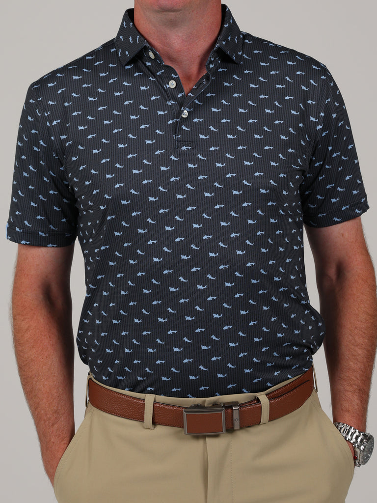 Man wearing a navy polo shirt with light blue shark pattern, beige pants, and a brown belt. He stands with hands in pockets against a neutral background.
