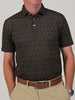 Man wearing a dark, football print polo shirt with small yellow paisley designs, beige pants, and a brown belt. He stands casually with his hands in his pockets.