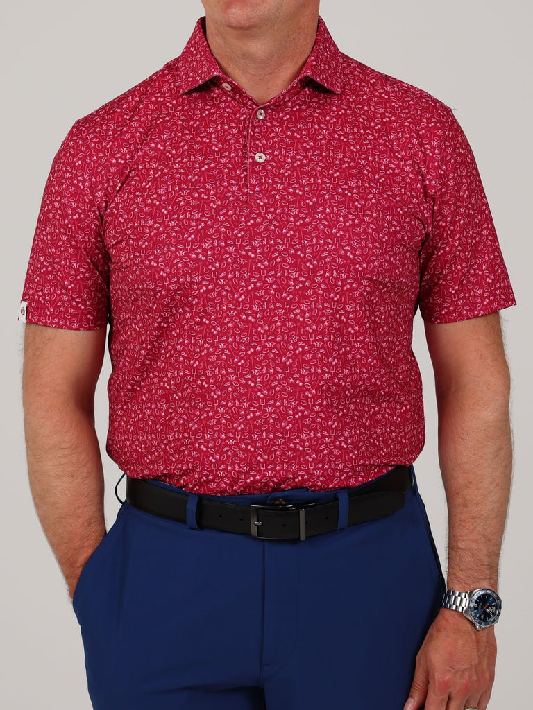 A man is wearing a red football patterned short-sleeve polo shirt with navy blue pants. He has a black belt and a silver watch.