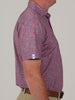 A man in a colorful, camoflauge patterned short-sleeve shirt with shades of red and blue stands in profile against a plain background. He's wearing tan pants.