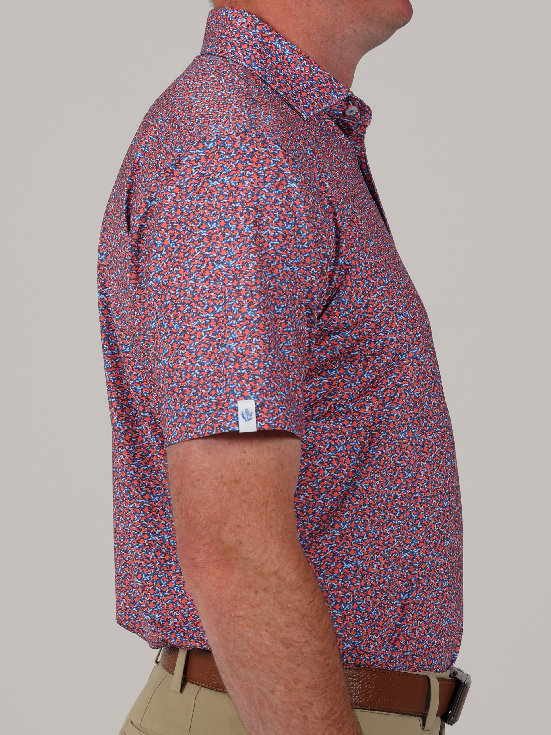 A man in a colorful, camoflauge patterned short-sleeve shirt with shades of red and blue stands in profile against a plain background. He's wearing tan pants.