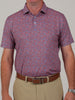 A man is wearing a red and blue camoflauge patterned polo shirt with a collar, paired with beige pants and a brown belt. The background is plain gray.