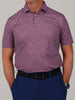 Man wearing a red and blue camoflauge patterned polo shirt with a causal stance. He has his hands in his pockets and wears a black belt with blue pants.