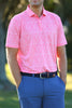 A man stands with his hands in his pockets, wearing a pink patterned polo shirt and blue trousers in a blurred outdoor setting.