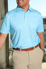 A man in a light blue polo shirt with white polka dots and khaki pants smiles while standing in a well-lit indoor space with a view.