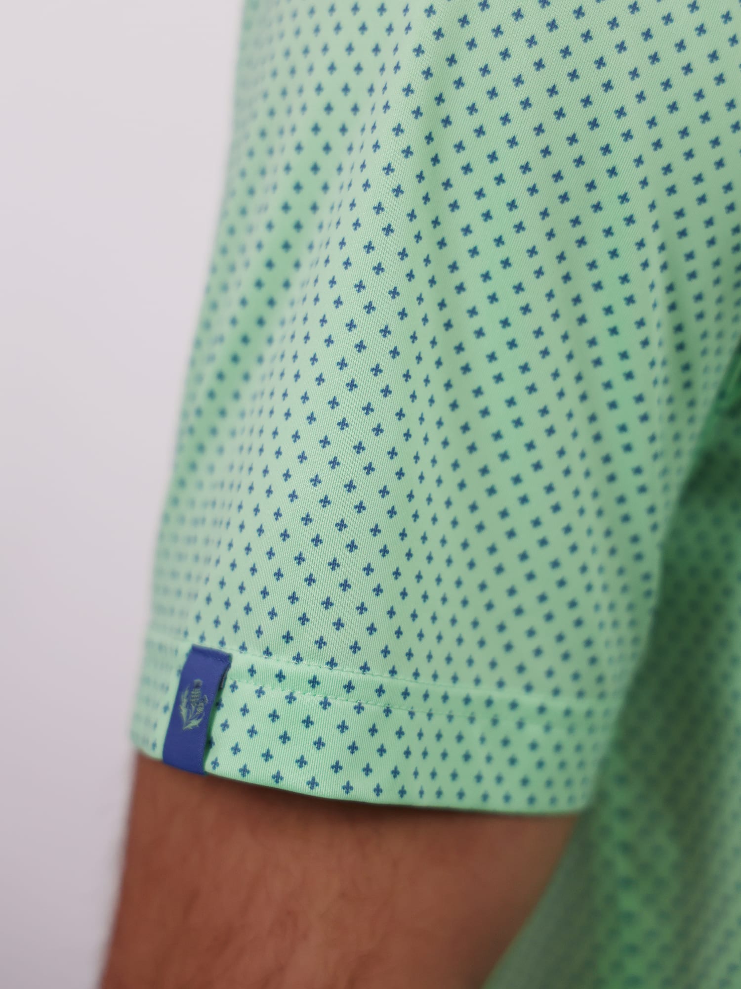 Close-up of a green shirt sleeve with a small fleur de lis pattern. A blue tag is visible near the hem