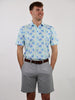 Man in a floral print shirt and gray shorts stands against a plain white background, hands in pockets, smiling confidently