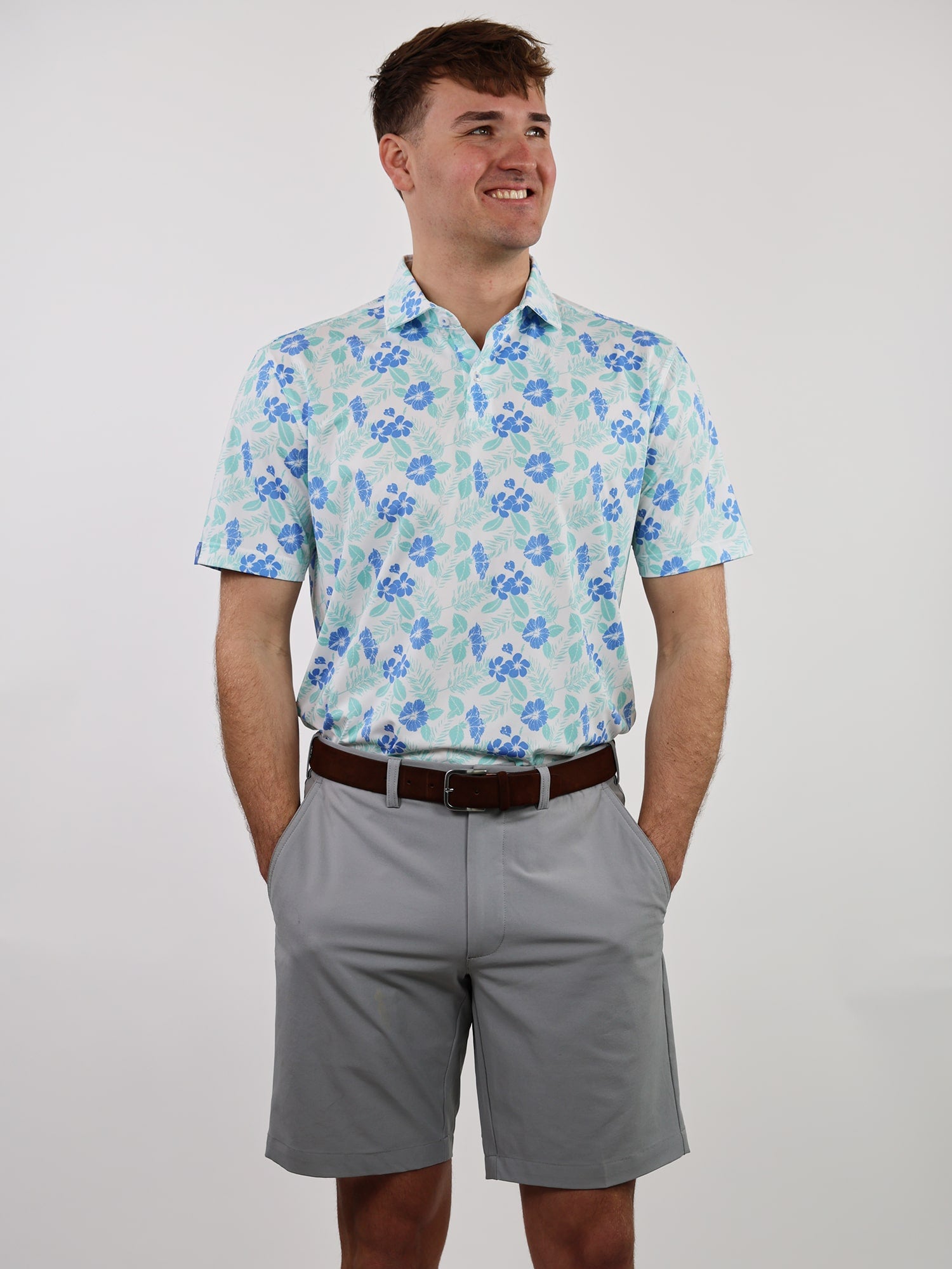 Man in a floral print shirt and gray shorts stands against a plain white background, hands in pockets, smiling confidently