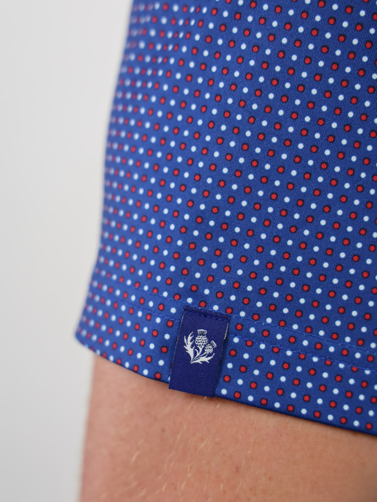Close-up of blue fabric with red and white polka dots, featuring a small blue tag with a white thistle emblem at the hem