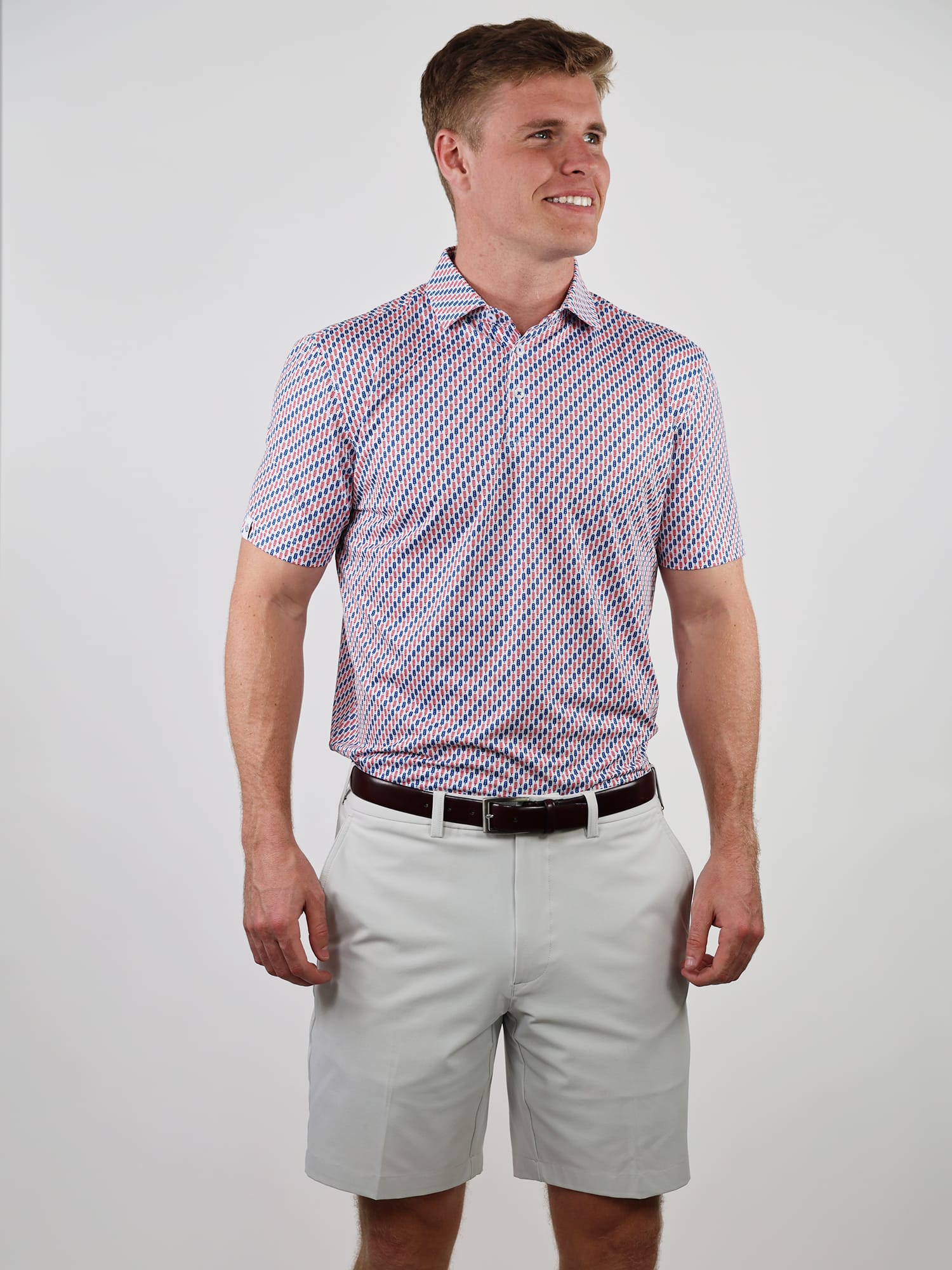 A man in a patterned polo shirt and light gray shorts stands smiling against a plain background