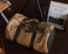 A vintage-style tan duffel bag with green accents rests on a dark wooden floor. Behind it, a book titled "Great Donald Ross Golf Courses" is visible.