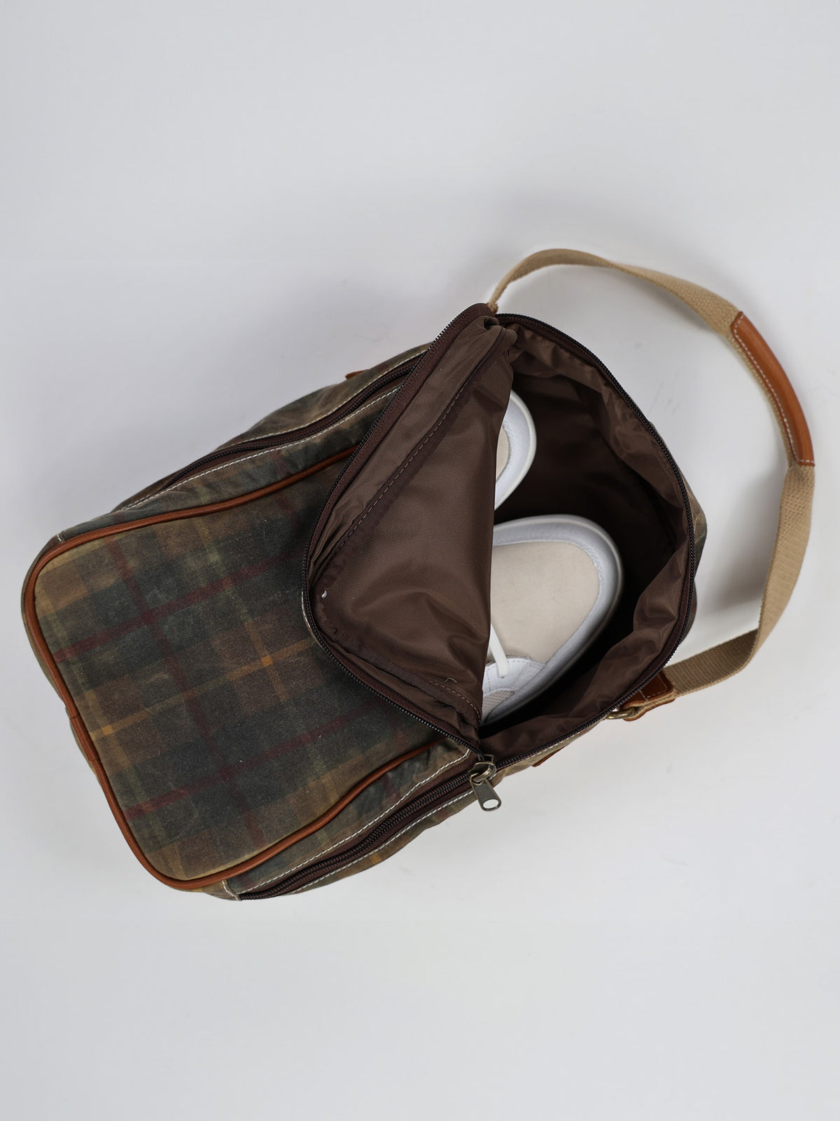 Plaid bag with a partially open zipper reveals white sneakers inside. The bag features tan handles and a brown interior lining.