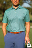 A man in a teal patterned polo shirt and blue pants holds a white golf towel with a logo in his right hand while standing with his left hand in his pocket.