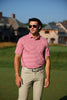 A man wearing a red and white striped polo shirt and khaki trousers stands on a manicured golf course with historic stone buildings in the background.