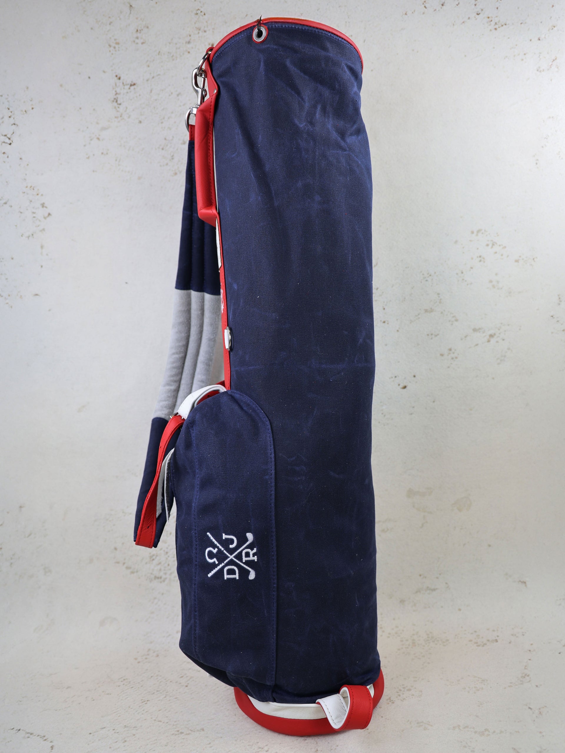 Navy canvas golf bag with red trim and embroidered crest logo displayed vertically against a white wall.