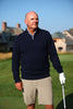 A mature man stands on a golf course wearing a navy quarter-zip sweater, khaki shorts, and white golf glove while holding a golf club.