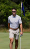 A man in a light blue patterned polo shirt and khaki shorts stands on a golf green holding the flagstick.