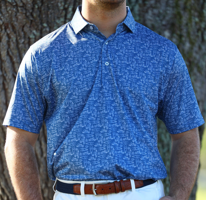 A person is wearing a blue patterned shirt with a collar and a brown belt, standing against a blurred tree background.