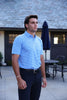 Man wearing a light blue striped polo shirt and navy trousers stands on a patio outside a modern home.