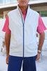 A man is standing and wearing a sleeveless white vest over a pink patterned shirt with blue shorts.
