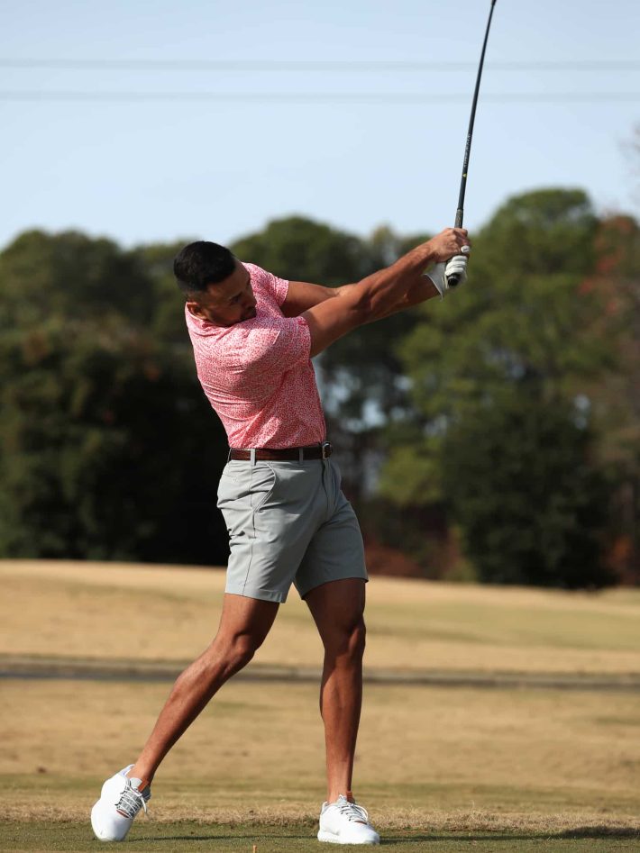 Man swinging a golf club wearing a men's tonal clovers polo in watermelon color.