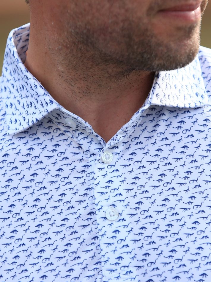 Close-up on the placket of a men's dinosaur print polo in white and navy.